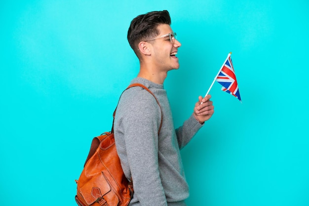 Jeune homme caucasien tenant un drapeau du Royaume-Uni isolé sur fond bleu en riant en position latérale