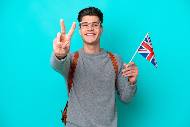 Jeune homme caucasien tenant un drapeau du Royaume-Uni isolé sur fond bleu souriant et montrant le signe de la victoire