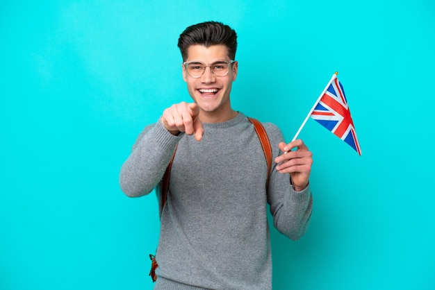 Jeune homme caucasien tenant un drapeau du Royaume-Uni isolé sur fond bleu surpris et pointant vers l'avant