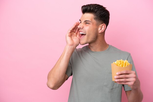 Jeune homme caucasien tenant des frites frites isolées sur fond rose criant avec la bouche grande ouverte sur le côté