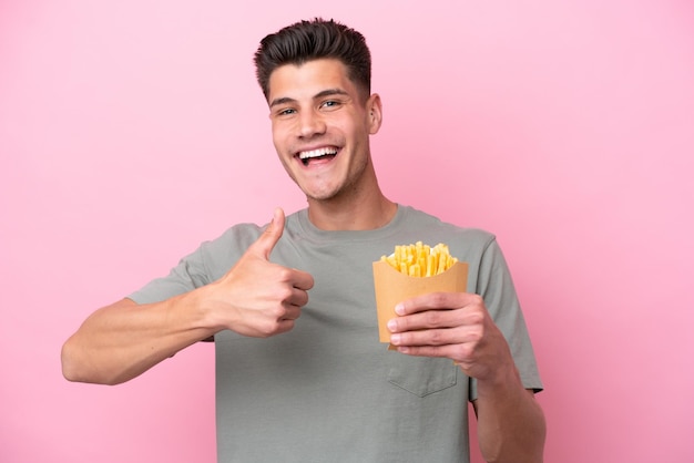 Jeune homme caucasien tenant des frites frites isolées sur fond rose donnant un geste du pouce levé