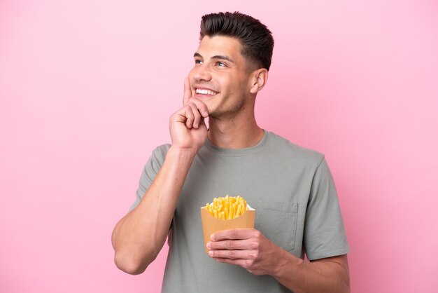 Jeune homme caucasien tenant des frites frites isolées sur fond rose en pensant à une idée tout en levant les yeux