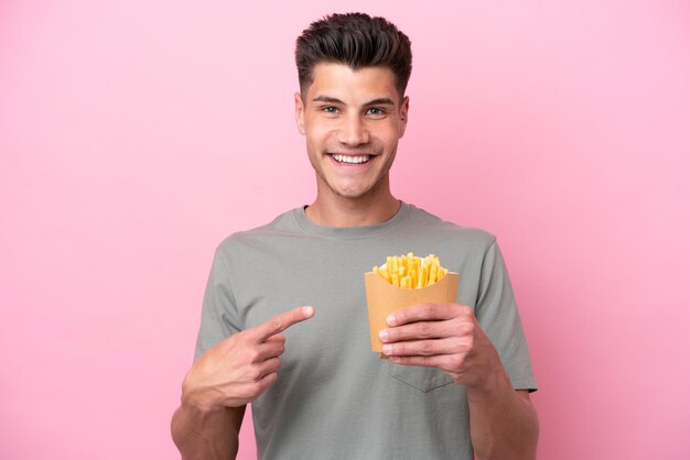 Jeune homme caucasien tenant des frites frites isolées sur fond rose pointant vers le côté pour présenter un produit