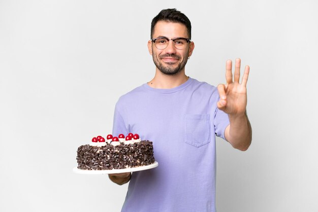 Jeune homme caucasien tenant un gâteau d'anniversaire isolé sur fond blanc heureux et comptant trois avec les doigts