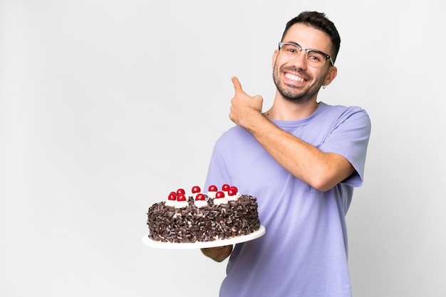 Jeune homme caucasien tenant un gâteau d'anniversaire isolé sur fond blanc pointant vers l'arrière