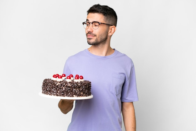 Jeune homme caucasien tenant un gâteau d'anniversaire isolé sur fond blanc regardant sur le côté