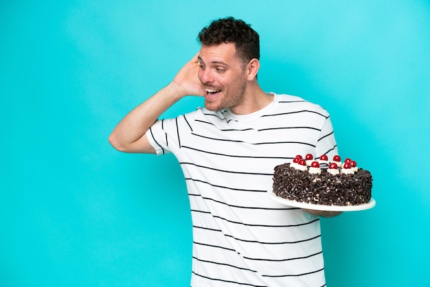 Jeune homme caucasien tenant un gâteau d'anniversaire isolé sur fond bleu écoutant quelque chose en mettant la main sur l'oreille