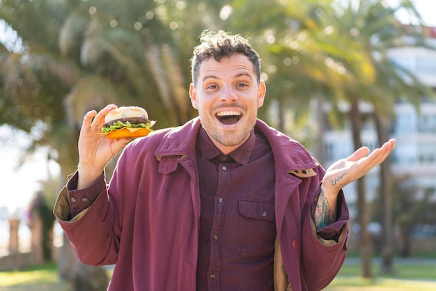 Jeune homme caucasien tenant un hamburger à l'extérieur avec une expression faciale choquée