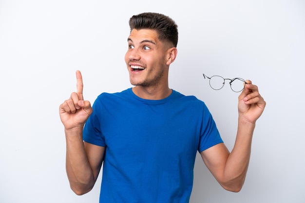 Jeune homme caucasien tenant des lunettes isolé sur fond blanc dans l'intention de réaliser la solution tout en levant un doigt