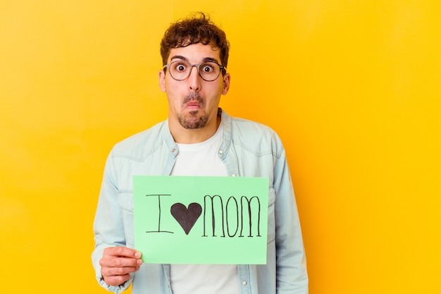 Jeune homme caucasien tenant une pancarte protéger notre planète hausse les épaules et les yeux ouverts confus.