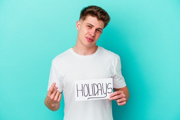 Jeune homme caucasien tenant une pancarte de vacances isolée sur mur bleu