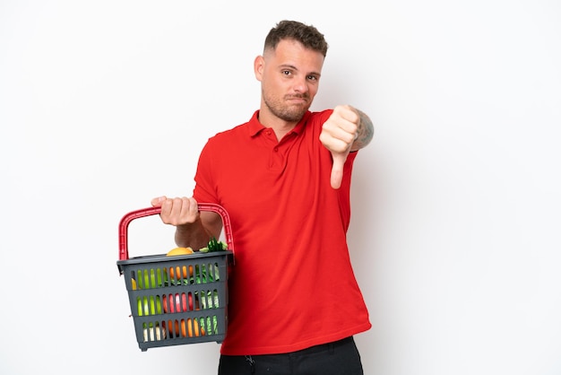 Jeune homme caucasien tenant un panier plein de nourriture isolé sur fond blanc montrant le pouce vers le bas avec une expression négative