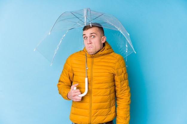 Jeune homme caucasien tenant un parapluie hausse les épaules et les yeux ouverts confus.