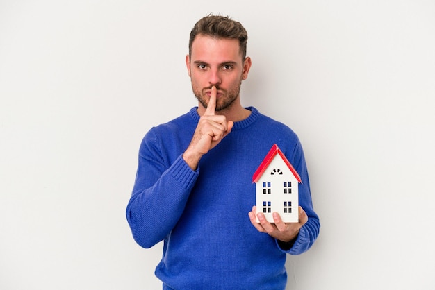 Jeune homme caucasien tenant une petite maison isolée sur fond blanc gardant un secret ou demandant le silence.