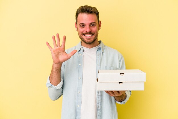 Jeune homme caucasien tenant une pizza isolée sur fond jaune souriant joyeux montrant le numéro cinq avec les doigts.