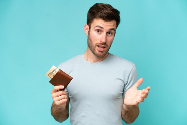 Jeune homme caucasien tenant un portefeuille isolé sur fond bleu avec une expression faciale choquée