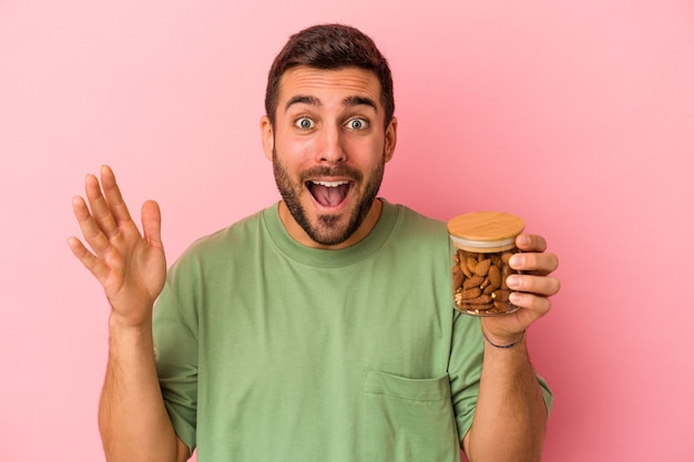 Jeune homme caucasien tenant un pot d'amande isolé sur un mur rose recevant une agréable surprise, excité et levant les mains.