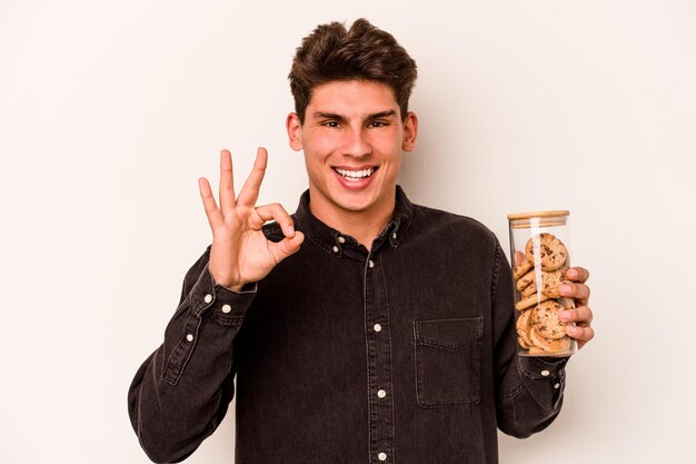 Jeune homme caucasien tenant un pot de biscuits isolé sur fond blanc joyeux et confiant montrant un geste ok