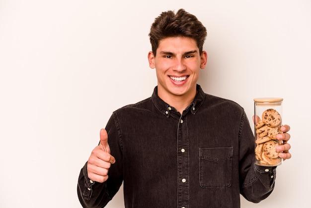 Jeune homme caucasien tenant un pot de biscuits isolé sur fond blanc souriant et levant le pouce vers le haut