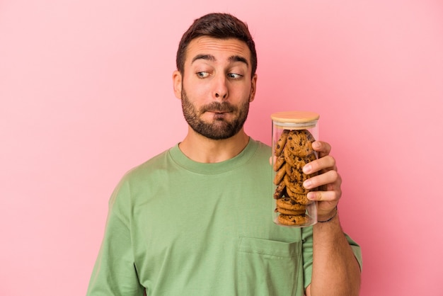 Jeune homme caucasien tenant un pot de biscuits isolé sur fond rose confus, se sent dubitatif et incertain.