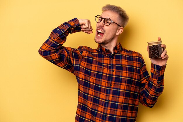 Jeune homme caucasien tenant un pot de café isolé sur fond jaune levant le poing après une victoire, concept gagnant.