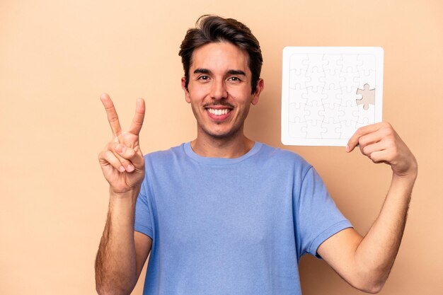 Jeune homme caucasien tenant un puzzle isolé sur fond beige montrant le numéro deux avec les doigts.