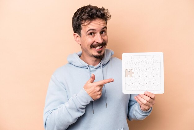 Jeune homme caucasien tenant un puzzle isolé sur fond beige souriant et pointant de côté, montrant quelque chose à l'espace vide.