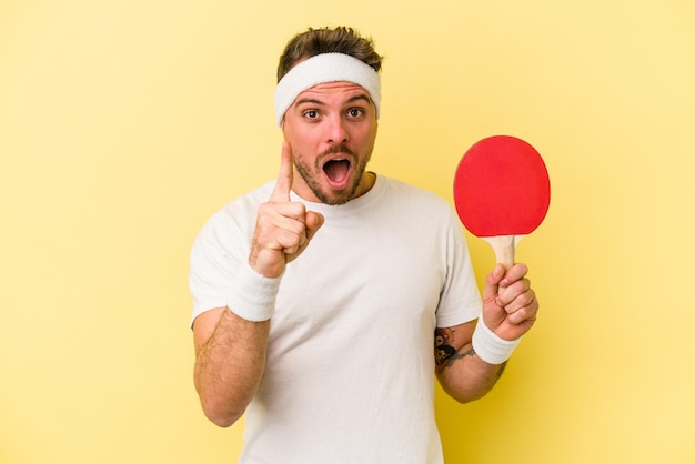 Jeune homme caucasien tenant une raquette de ping-pong isolée sur fond jaune ayant une idée, un concept d'inspiration.