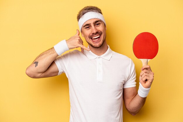 Jeune Homme Caucasien Tenant Des Raquettes De Ping-pong Isolées Sur Fond Jaune Montrant Un Geste D'appel De Téléphone Portable Avec Les Doigts.