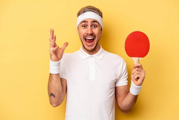 Jeune homme caucasien tenant des raquettes de ping-pong isolées sur fond jaune recevant une agréable surprise, excité et levant les mains.