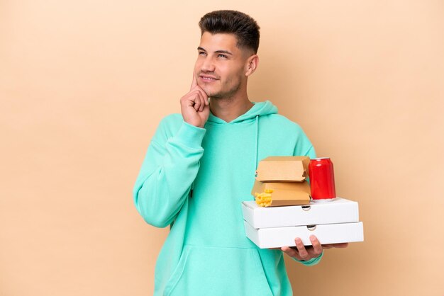 Jeune homme caucasien tenant de la restauration rapide isolé sur fond beige en pensant à une idée tout en levant les yeux