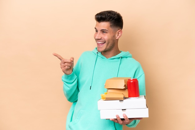 Jeune homme caucasien tenant de la restauration rapide isolé sur fond beige pointant le doigt sur le côté et présentant un produit