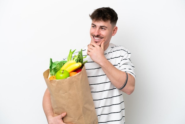 Jeune homme caucasien tenant un sac d'épicerie isolé sur fond blanc regardant sur le côté et souriant