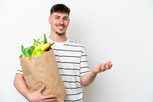 Jeune homme caucasien tenant un sac d'épicerie isolé sur fond blanc tendant les mains sur le côté pour inviter à venir