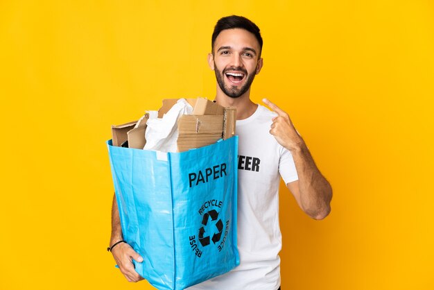 Jeune homme caucasien tenant un sac de recyclage plein de papier à recycler isolé sur fond blanc donnant un geste de pouce en l'air