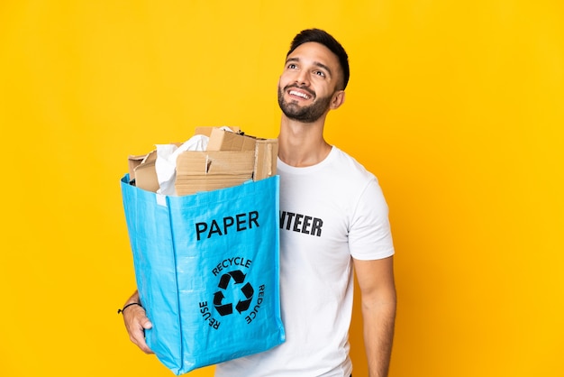 Jeune homme caucasien tenant un sac de recyclage plein de papier à recycler isolé sur fond blanc en pensant à une idée tout en levant les yeux