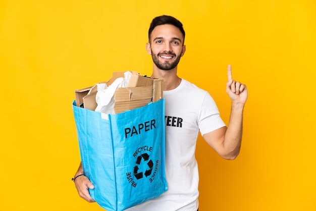 Jeune homme caucasien tenant un sac de recyclage plein de papier à recycler isolé sur fond blanc pointant vers le haut une excellente idée