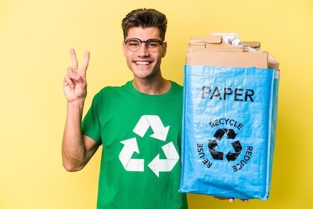 Jeune homme caucasien tenant un sac de recyclage plein de papier à recycler isolé sur fond jaune montrant le numéro deux avec les doigts.