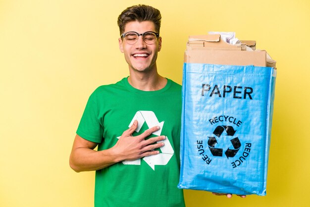 Jeune homme caucasien tenant un sac de recyclage plein de papier à recycler isolé sur fond jaune rit bruyamment en gardant la main sur la poitrine.
