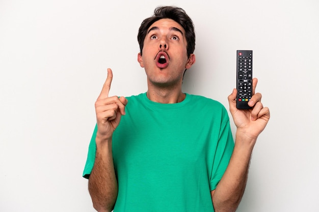 Jeune homme caucasien tenant une télécommande de télévision isolée sur fond blanc pointant vers le haut avec la bouche ouverte