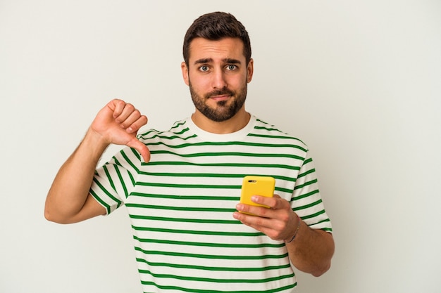 Jeune homme caucasien tenant un téléphone mobile isolé sur un mur blanc montrant un geste d'aversion, les pouces vers le bas. Concept de désaccord.