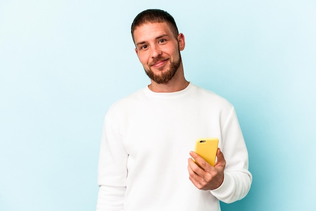 Jeune homme caucasien tenant un téléphone portable isolé sur fond bleu heureux, souriant et joyeux.