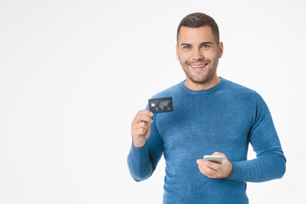 Un jeune homme caucasien en tenue décontractée tenant une carte de crédit et un smartphone isolé sur un fond blanc
