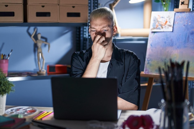 Jeune homme caucasien utilisant un ordinateur portable la nuit dans un studio d'art sentant quelque chose de puant et dégoûtant, une odeur intolérable, retenant son souffle avec les doigts sur le nez. mauvaise odeur