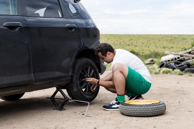 Jeune homme changeant le pneu crevé sur sa voiture en desserrant les écrous avec une clé de roue avant de soulever le véhicule