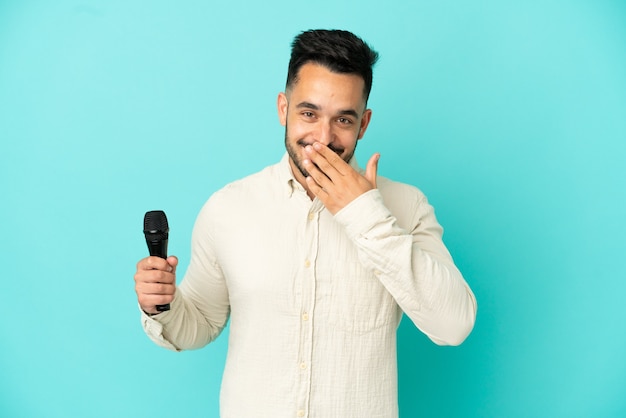 Jeune homme chanteur caucasien isolé sur fond bleu heureux et souriant couvrant la bouche avec la main