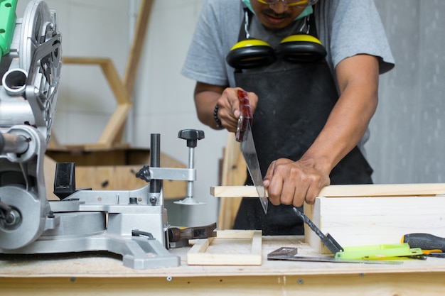 Jeune homme charpentier coupant le bois à l'aide d'une scie à main sur sa table d'atelier portant un équipement de sécurité