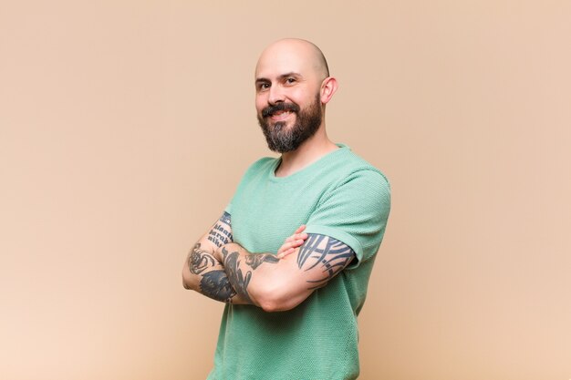 Jeune homme chauve et barbu souriant à la caméra avec les bras croisés et une expression heureuse, confiante et satisfaite, vue latérale