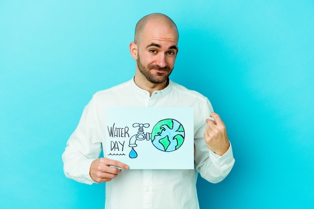 Jeune homme chauve caucasien célébrant la journée mondiale de l'eau isolée sur bleu pointant avec le doigt sur vous comme si vous invitiez à vous rapprocher.