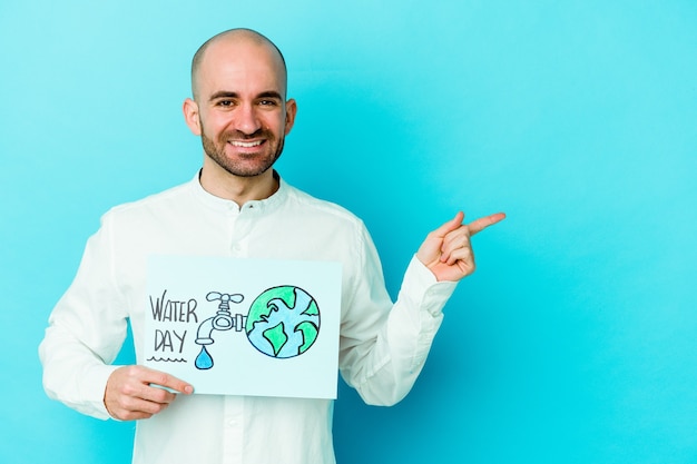 Jeune homme chauve caucasien célébrant la journée mondiale de l'eau isolée sur fond bleu souriant et pointant de côté